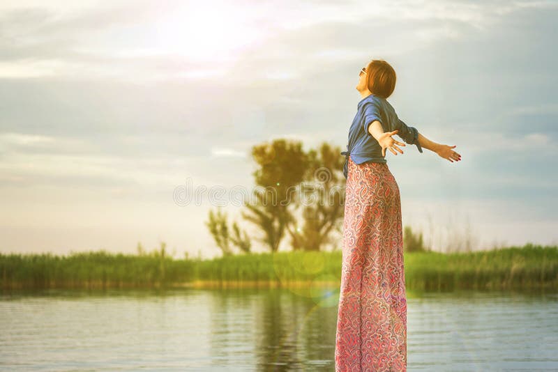 Beautiful young red haired girl in colorful long sarafan dress sitting on a wooden pier on a river or lake. Green bulrush and tree stock image Beautiful young red haired girl in colorful long sarafan dress sitting on a wooden pier on a river or lake. Green bulrush and tree stock image