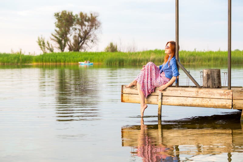Beautiful young red woman sitting on a wooden pier in a colorful long sarafan dress and putting her leg in a water royalty free stock photo Beautiful young red woman sitting on a wooden pier in a colorful long sarafan dress and putting her leg in a water royalty free stock photo
