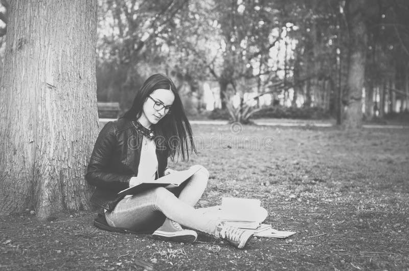 Beautiful young school or college girl with long hair and eye glasses sitting on the ground in the park reading the books and stud