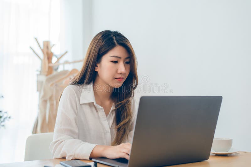Beautiful young smiling Asian woman working on laptop while at home in office work space.