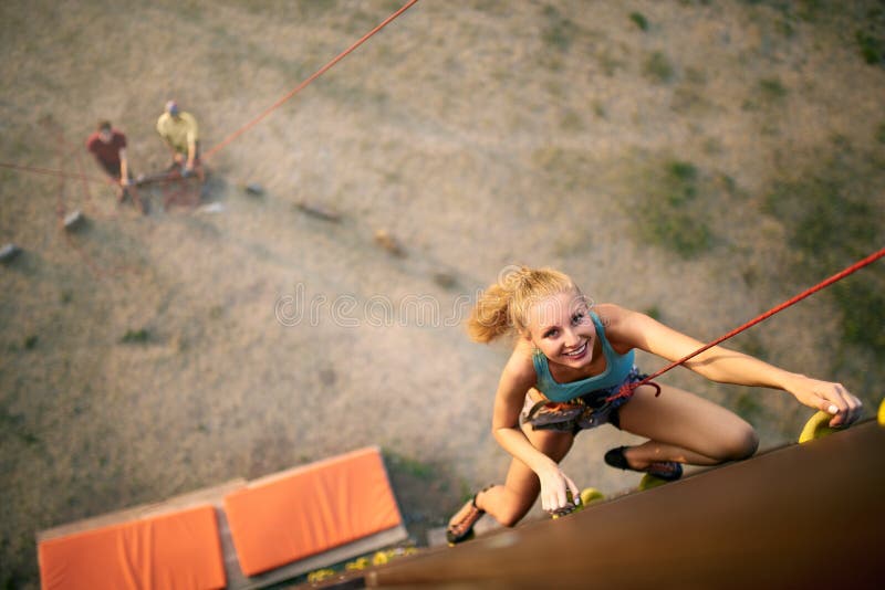 Beautiful young strong woman climbing on rock artificial wall in summer, top view. Climber insured on belaying harness