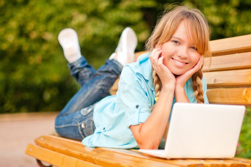 Beautiful young student girl lying on bench with laptop, her hands under the chin, smiling and looking into the camera