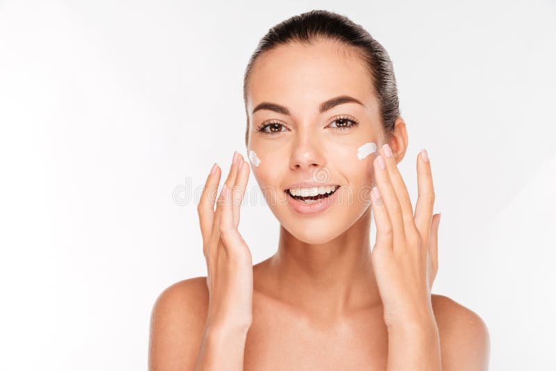 Beautiful young woman applying cosmetic cream treatment on her face isolated on white background