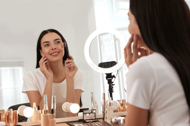 Beautiful young woman applying makeup at table with mirror and ring lamp