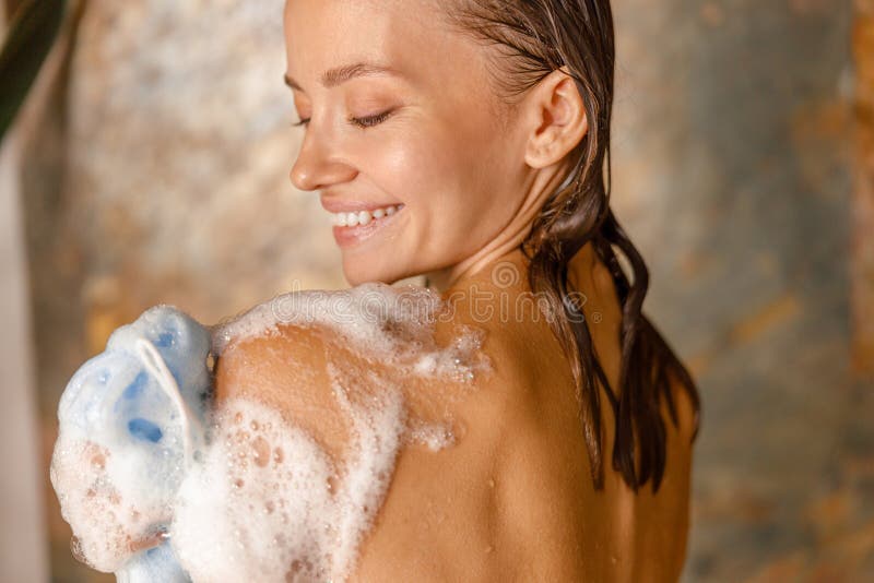 Beautiful young woman applying shower gel using loofah sponge while taking shower