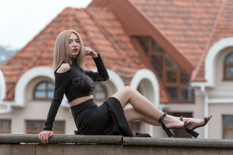 Beautiful young woman with blonde hair wears black clothes long skirt and heels. Stylish girl outside. Old building on background