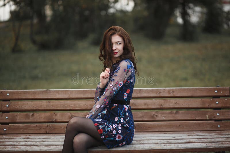 Beautiful young woman with brown curly hair sitting on wooden bench in autumn park. She keeping her hair and smiling