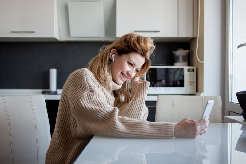 Beautiful young woman in a cozy sweater is sitting in the kitchen using a video call in the messenger.