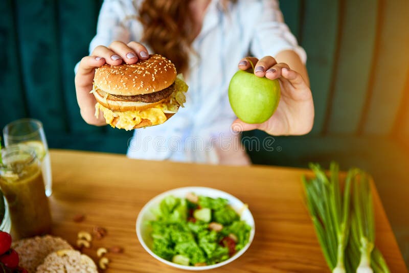 Beautiful young woman decides eating hamburger or apple in kitchen. Cheap junk food vs healthy diet royalty free stock image Beautiful young woman decides eating hamburger or apple in kitchen. Cheap junk food vs healthy diet royalty free stock image