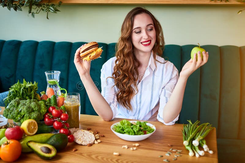 Beautiful young woman decides eating hamburger or apple in kitchen. Cheap junk food vs healthy diet