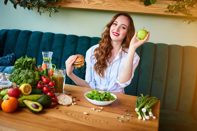 Beautiful young woman decides eating hamburger or apple in kitchen. Cheap junk food vs healthy diet stock images Beautiful young woman decides eating hamburger or apple in kitchen. Cheap junk food vs healthy diet stock images