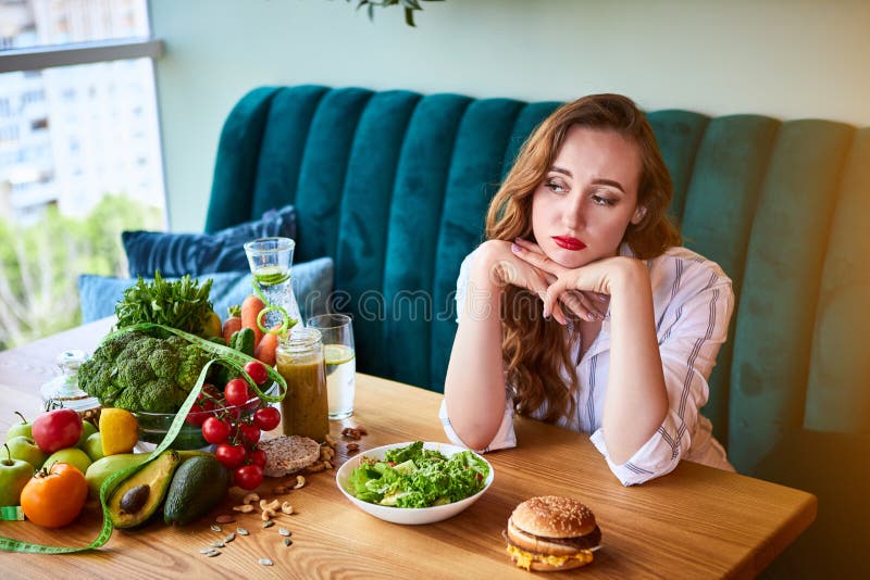 Beautiful young woman decides eating hamburger or vegetables  in kitchen. Cheap junk food vs healthy diet