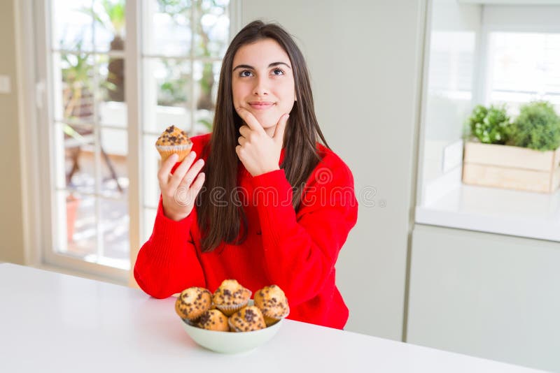 Beautiful young woman eating chocolate chips muffins serious face thinking about question, very confused idea