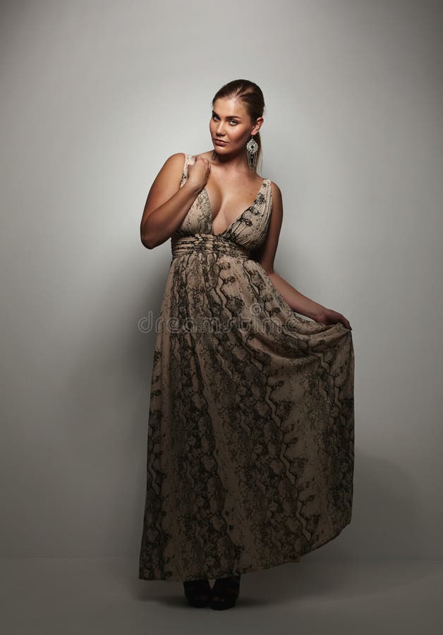 Beautiful young woman in a evening dress standing on grey background looking at camera. Plus size caucasian female model posing in evening gown.