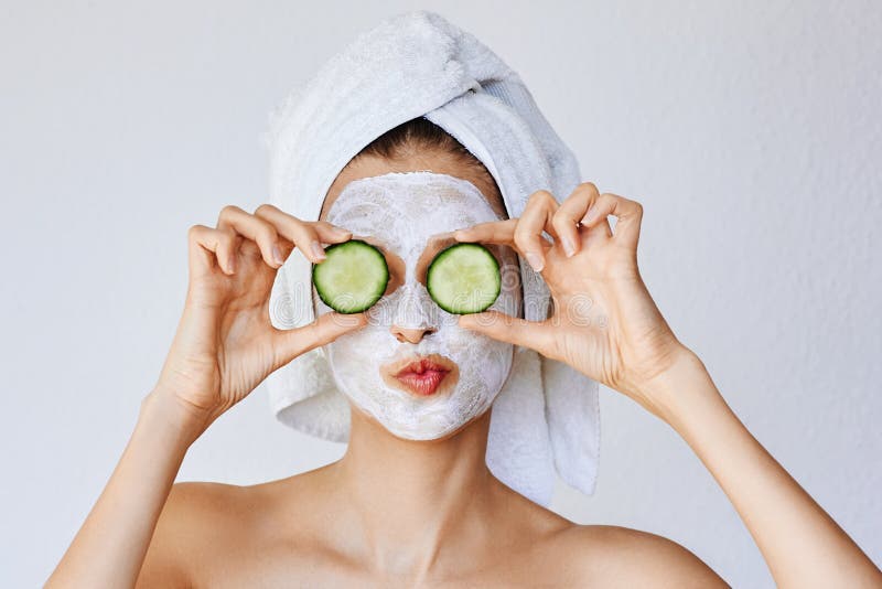 Beautiful young woman with facial mask on her face holding slices of cucumber. Skin care and treatment, spa, natural beauty and