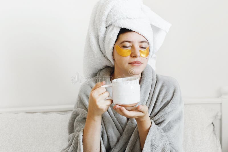 Beautiful young woman with golden eye patches and in bathrobe holding cup of coffee and lying in bed, enjoying morning routine. royalty free stock photo Beautiful young woman with golden eye patches and in bathrobe holding cup of coffee and lying in bed, enjoying morning routine. royalty free stock photo