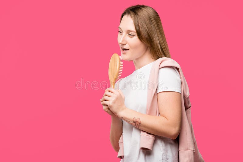 beautiful young woman holding hairbrush and singing isolated on pink