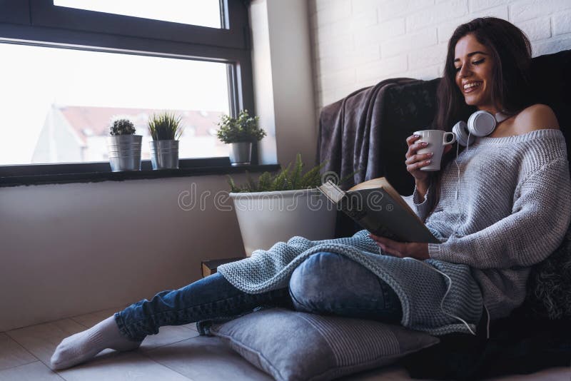 Beautiful young woman at home drinking coffee reading a book