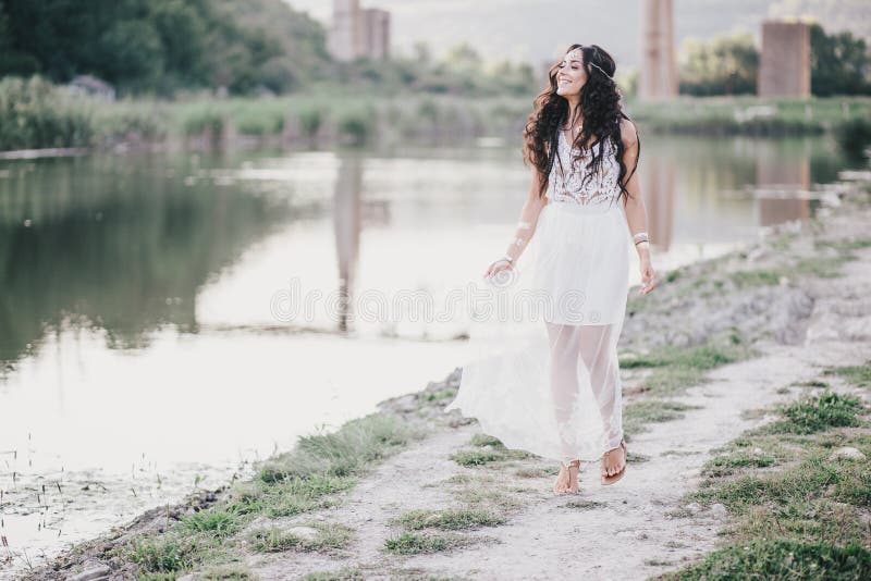 Beautiful young woman with long curly hair dressed in boho style dress posing near lake