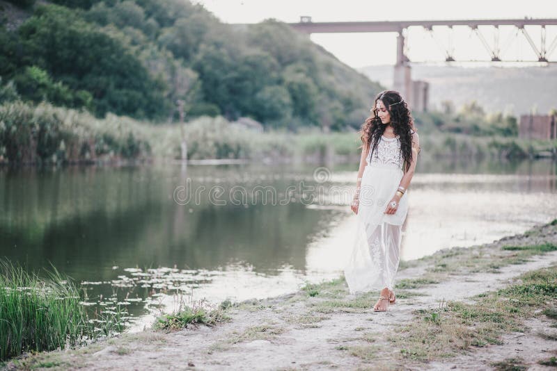 Beautiful young woman with long curly hair dressed in boho style dress posing near lake