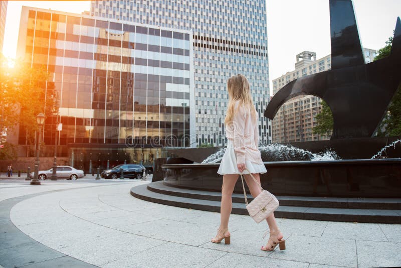 Beautiful young woman with long legs walking on city street wearing short skirt and pink t-shirt and holding a bag