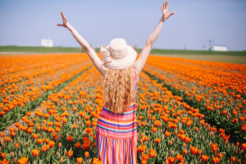 Beautiful young woman with long red hair wearing a striped dress and straw hat standing by the back on colorful tulip field