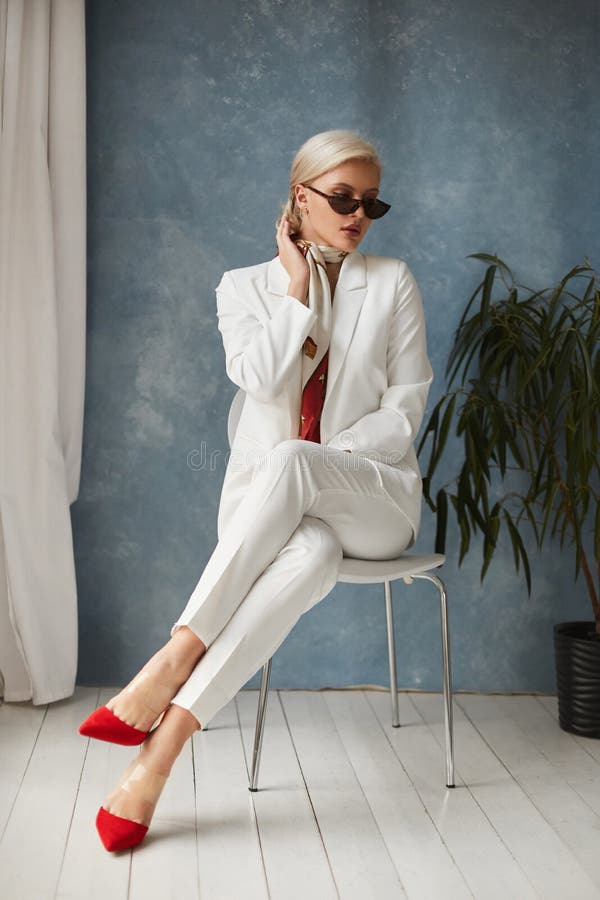 Beautiful young woman with perfect blond hair in elegant white suit and red shoes posing in studio. Fashionable blonde