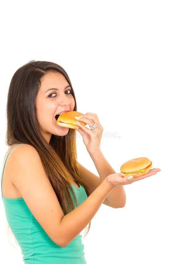 Beautiful young woman posing for camera eating hamburger while holding another one in other hand, smiling happily, white royalty free stock photography