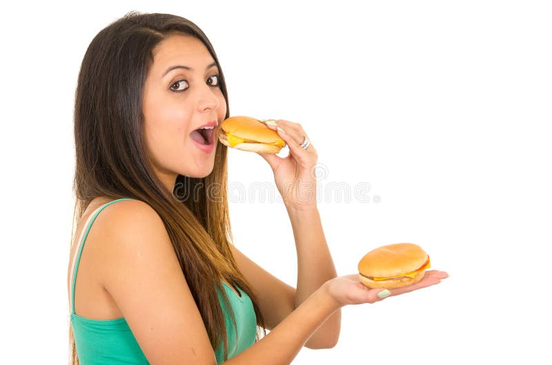 Beautiful young woman posing for camera eating hamburger while holding another one in other hand, smiling happily, white royalty free stock photos