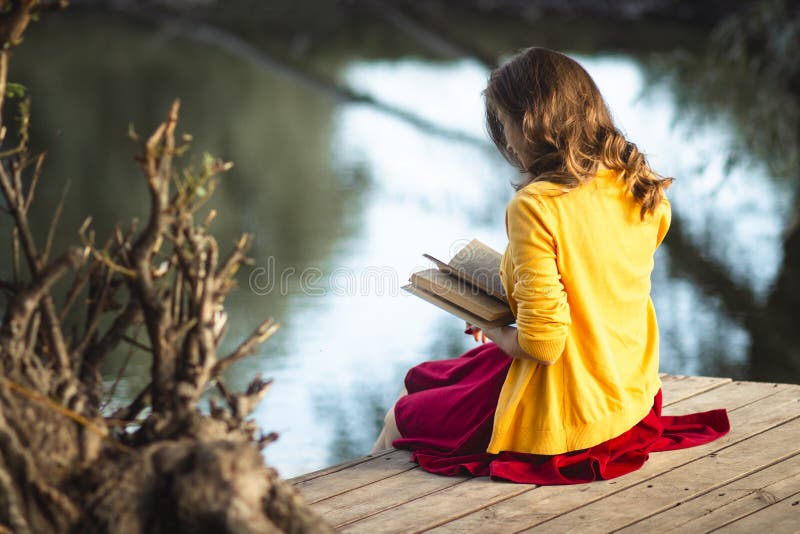 Beautiful young woman on the river banks sitting on a wooden platform with a book, teenager girl reading fiction literature on