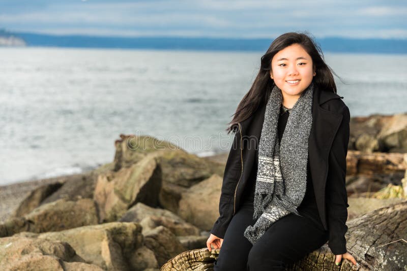 Beautiful young Asian American teenage girl with long black hair and dressed in a black outfit and grey scarf sits on a large piece of driftwood on a rocky beach. Ocean and sky in background. Horizontal. Copy space. Beautiful young Asian American teenage girl with long black hair and dressed in a black outfit and grey scarf sits on a large piece of driftwood on a rocky beach. Ocean and sky in background. Horizontal. Copy space.