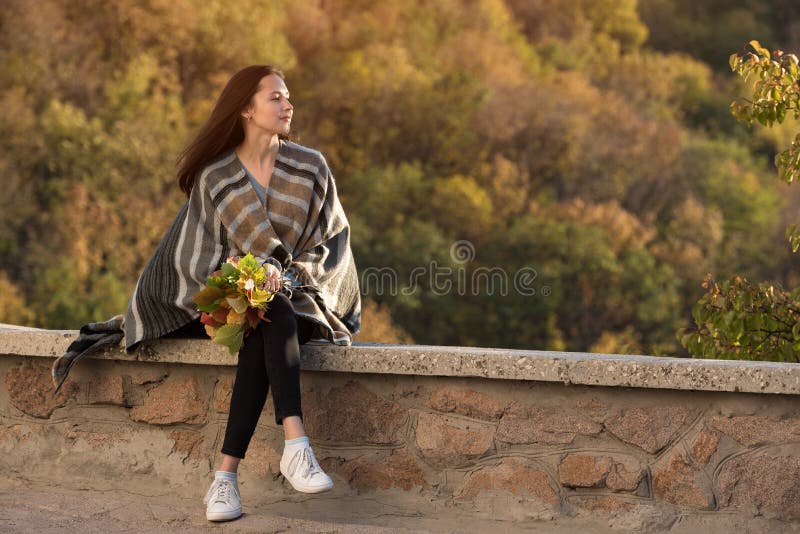 Beautiful young woman sitting on the roadside on nature background and thinking about life. Bouquet of autumn leaves in her hands