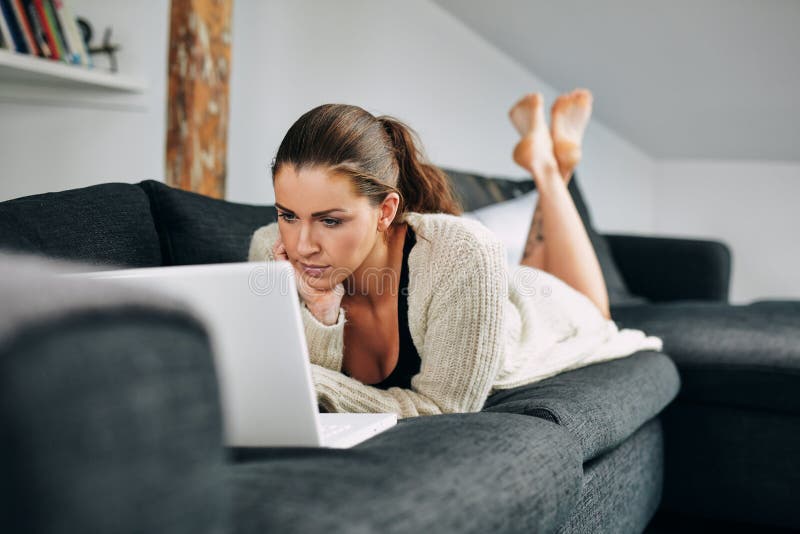 Portrait of beautiful young woman working on laptop while lying on sofa. Female using laptop at home. Portrait of beautiful young woman working on laptop while lying on sofa. Female using laptop at home.