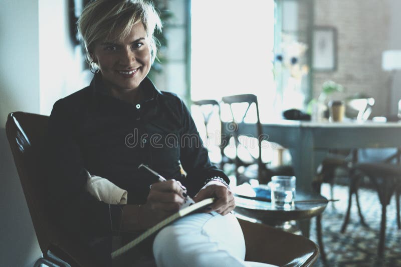 Beautiful young woman writing something in note pad while sitting on armchair at living room.Charming female studying stock photo Beautiful young woman writing something in note pad while sitting on armchair at living room.Charming female studying stock photo