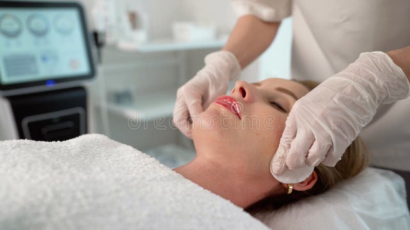 Beautiker som applicerar kräm på kvinnans ansikte vid hudbehandling. Beautiker som massakrerar en kund medan hon slappnar av på en bår i en modern skönhetssalon och får en föryngrande hudbehandling