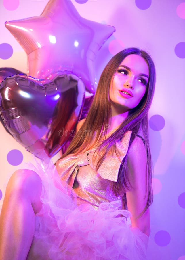 Beauty fashion model party Valentine`s Day girl with heart shaped air balloons, sitting on chair, laughing in neon light Holiday celebration. Beautiful young pin up brunette woman full length portrait