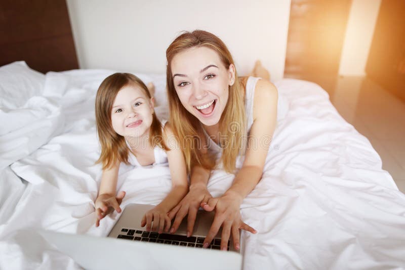 Beauty mother hugs little daughter with laptop laying at white bed with sunshine