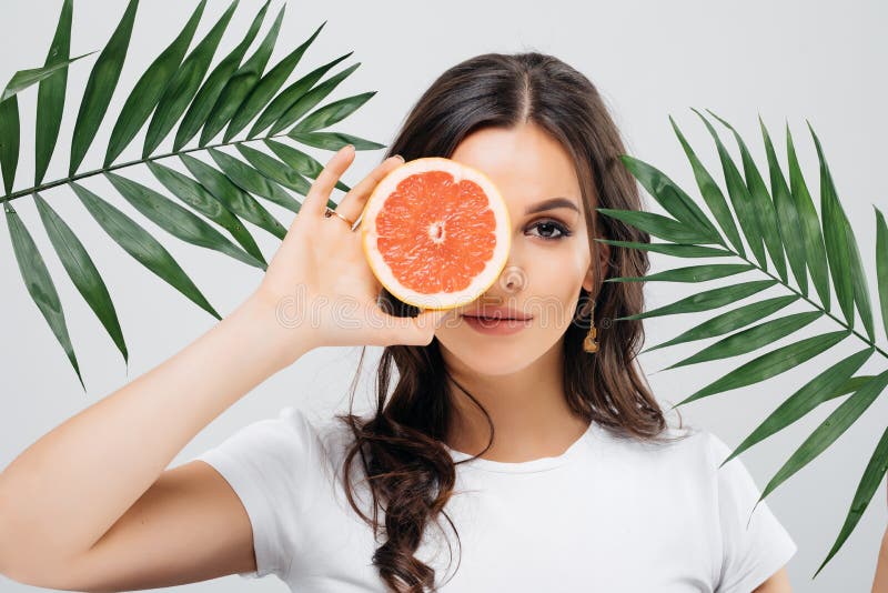 Portrait of an excited woman with brunette hair posing with green leaves and holding sliced grapefruit isolated over white backgro