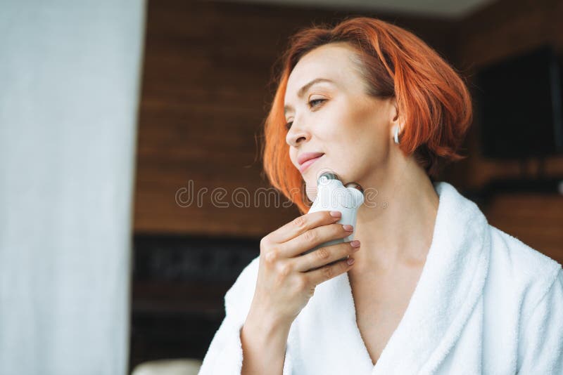 Beauty portrait of smiling woman 35 year in white bathrobe with clean fresh face and hands with red hair doing fasial massage with