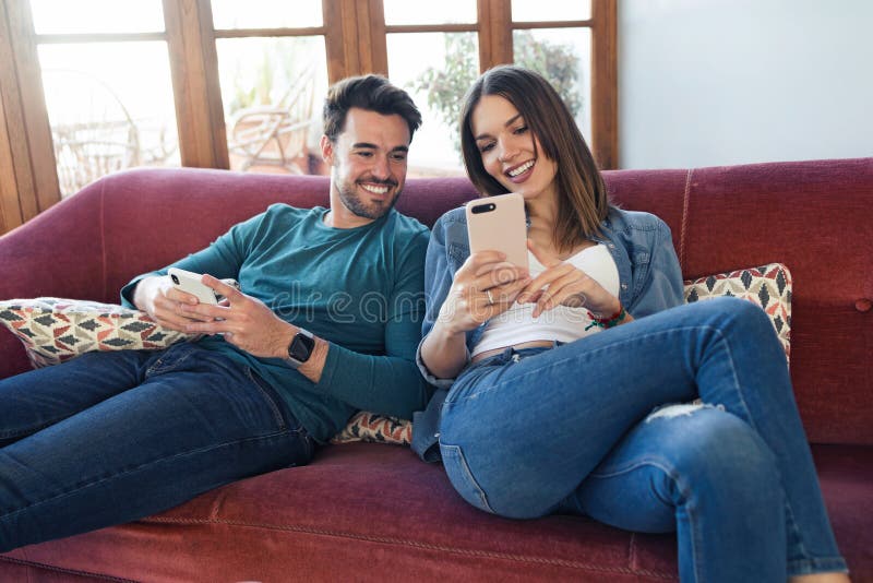 Beaux jeunes couples employant ils t?l?phone portable tout en se reposant sur le sofa ? la maison photo stock Beaux jeunes couples employant ils t?l?phone portable tout en se reposant sur le sofa ? la maison photo stock