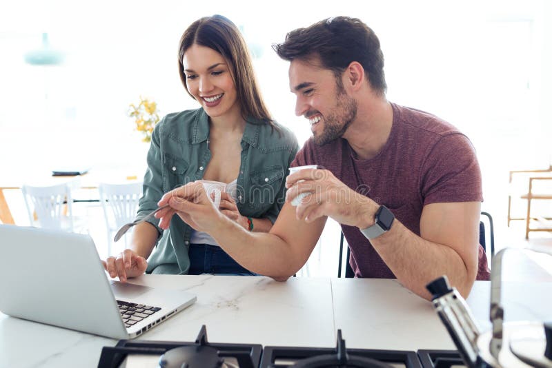 Beaux beaux jeunes couples utilisant leur ordinateur portable et petit d?jeuner de avoir dans la cuisine ? la maison image stock Beaux beaux jeunes couples utilisant leur ordinateur portable et petit d?jeuner de avoir dans la cuisine ? la maison image stock