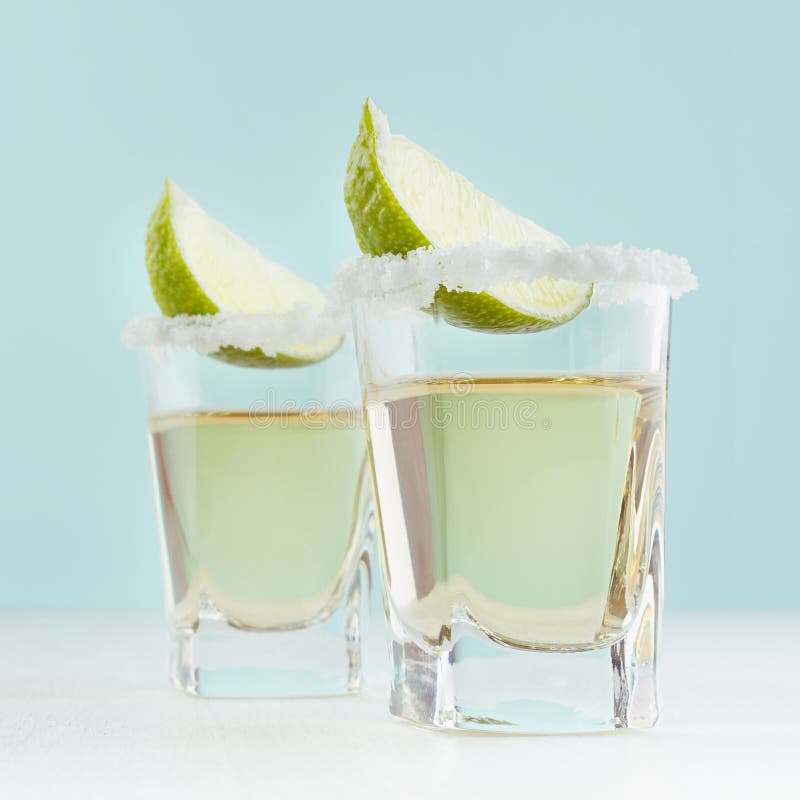 Bebida de oro mexicana tradicional del alcohol del tequila con el borde de la cal y de la sal en vaso de medida en el fondo azul