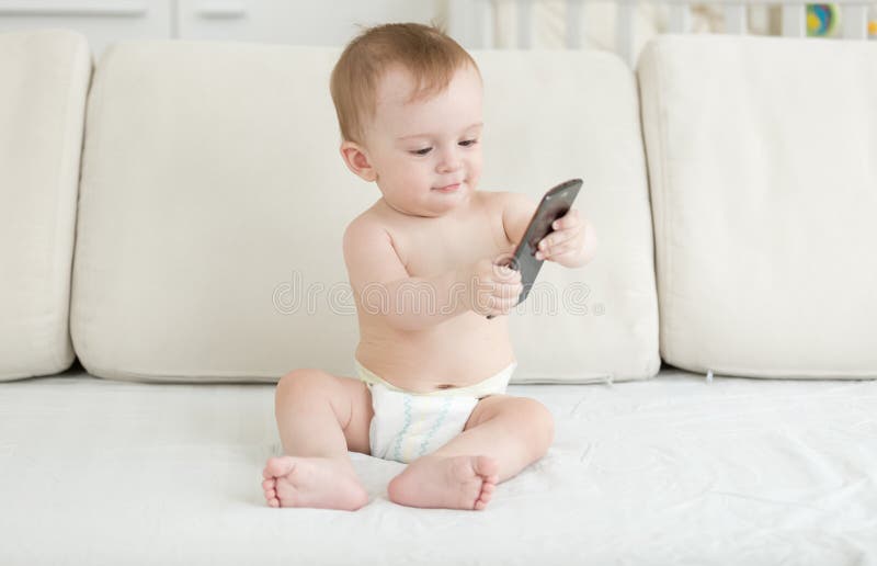 Bebé adorable en los pañales que se sientan en el sofá y que usan smartphone foto de archivo libre de regalías Bebé adorable en los pañales que se sientan en el sofá y que usan smartphone foto de archivo libre de regalías