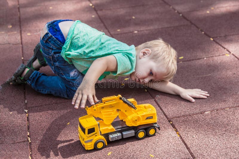 Little boy with blue eyes laying on the cover of children playground, playing with toy - yellow excavator. Wearing aquamarine t-short and jeans, with blonde hair. Outdoors, sunny day, copy space. Little boy with blue eyes laying on the cover of children playground, playing with toy - yellow excavator. Wearing aquamarine t-short and jeans, with blonde hair. Outdoors, sunny day, copy space