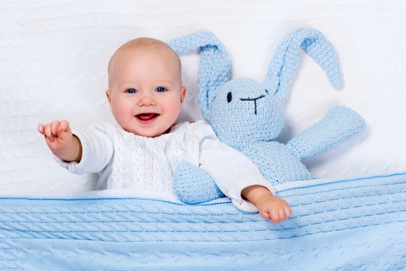 Funny little baby wearing a warm knitted jacket playing with toy bunny relaxing on white cable knit blanket in sunny nursery. Kids winter clothing and bedding. Hand made toys and textile for children. Funny little baby wearing a warm knitted jacket playing with toy bunny relaxing on white cable knit blanket in sunny nursery. Kids winter clothing and bedding. Hand made toys and textile for children.