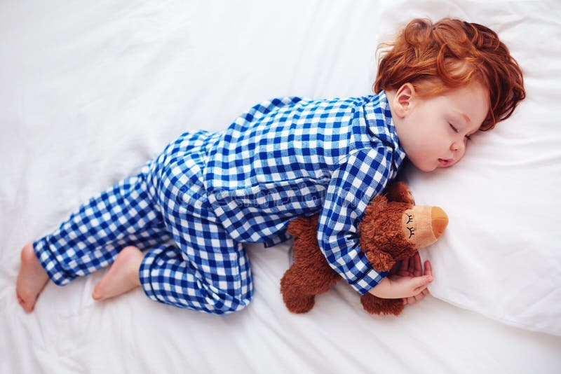 Bebê adorável da criança do ruivo que dorme com o brinquedo do luxuoso em pijamas da flanela fotos de stock royalty free Bebê adorável da criança do ruivo que dorme com o brinquedo do luxuoso em pijamas da flanela fotos de stock royalty free