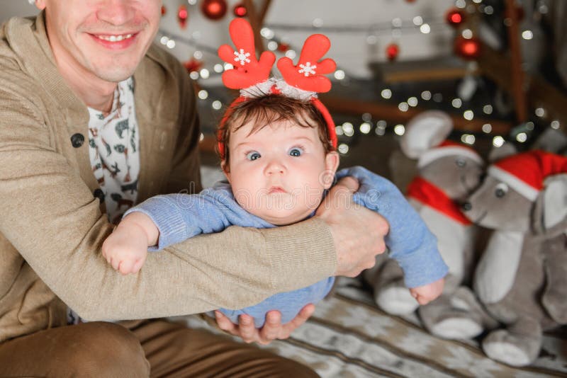 Bebê em chifres de veados vermelhos feliz Ano Novo um fato para crianças Natal O pai segura nos braços e treme de um lado para o fotos de stock royalty free Bebê em chifres de veados vermelhos feliz Ano Novo um fato para crianças Natal O pai segura nos braços e treme de um lado para o fotos de stock royalty free
