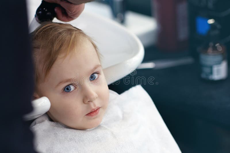 Bebê louro bonito assustado sério e pequeno com olhos azuis em uma barbearia que tem a cabeça de lavagem pelo cabeleireiro imagens de stock Bebê louro bonito assustado sério e pequeno com olhos azuis em uma barbearia que tem a cabeça de lavagem pelo cabeleireiro imagens de stock