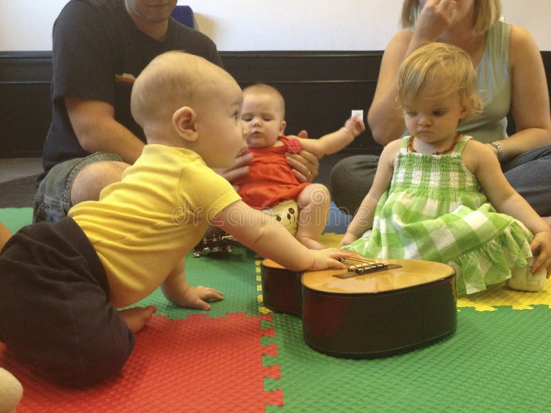 Babies in a music class are gathered around a guitar which has been placed on the floor for them to play with it. The parents watch from the background. Babies in a music class are gathered around a guitar which has been placed on the floor for them to play with it. The parents watch from the background