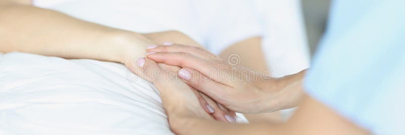 On bed lies a sick doctor sitting next to him and sympathetically holds hand